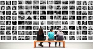 Three people seated in front of a wall of pictures of people's faces, with all faces communicating something.