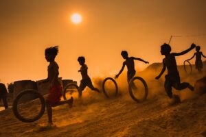 An image of children playing. They are in silhouette form, backlit by the setting sun. They are running downhill, rolling tyres as they go