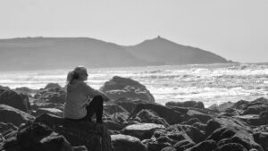 A woman sitting on some rocks beside the sea. The sun is shining and she is daydreaming.