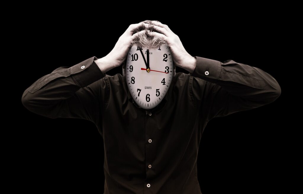 A picture of a person, their face replaced with a clock with is showing a time of 11:55. The person is holding their head, stressed out.