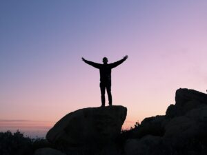 A person, in silhouette, celebrating their success as they stand on top of a rock with the sun about to rise in the background