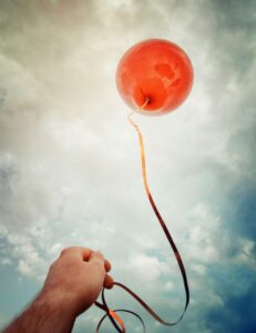 A person holding a single red balloon. Are they holding on or are they letting go? Is this you? What are you holding on to? Are you ready to let go?
