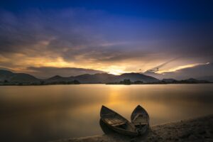 The sun rising over hills casting shadows and colour over a perfectly still lake. A couple of wooden boats sit on the shore. There is a peace and calmness about the picture.