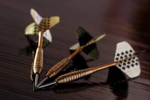 Three metal darts laying on a dark wooden surface. Their tips are touching, all focussed on the same point.