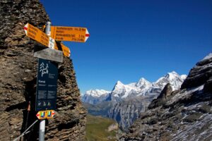 A signpost on a mountain pass, with mountains in the background, some of them with snow at the top. The signs are pointing to all the options.