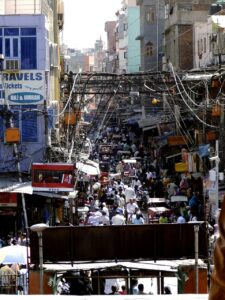 A chaotic street packed with people, buildings of varying shapes, sizes and colours lining the street, with masses of cables forming a tangled web overhead.