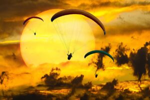 There are three paragliders flying through the air. They are backlit by a bright sun. They've taken a leap of faith. Can you?