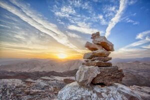 A carefully balanced pile of rocks on top of a mountain. The sun is rising in the distance, showing a vast landscape.