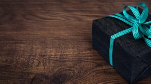 A gift wrapped in black textured paper with a bright blue ribbon tying it up. The package sits on a wooden table top. The package, like life, is full of surprises.
