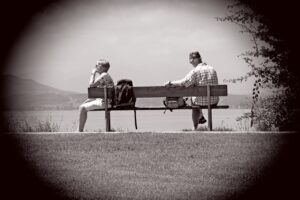 A black and white photo of a man and a woman sitting on a bench. they are beside a lake on a sunny day. The woman has her back to the man. There is no communication here.