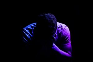 A person, head in hands, full of despair. They are partially lit by blue and purple lighting - it is a picture of gloom and despondency.