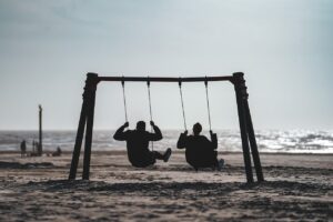 Two people on the beach, swinging on two swings. There are clear skies, the sea is calm, and the two people seem happy. Thet are in silhouette.