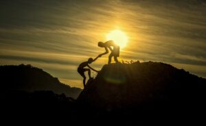 the sun is rising behind two people who are helping each other to climb what looks like a hill or rocky point. It is their mountain and their challenge.