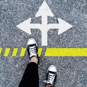 A person standing at a junction, with an arrow pointing to the left, to the right and straight on. We can only see the person's feet as they stop and decide which route leads to a better future