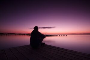 A person, in silhouette, is sitting beside a lake. The sun is about to rise and the sky is varying shades of pink, orange and purple. It is a picture of absolute calm.