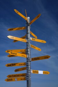 A sign post with a blue sky behind it. The post has multiple signs on it, pointing to different cities around the world.