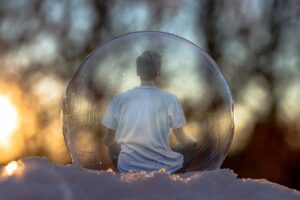 A person is sitting in a bubble which is resting on snow. In the out of focus background the sun is shining through trees. The person is contemplating life and how to get back on track.