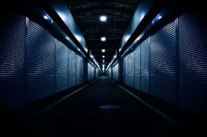 A dark alleyway, with closed metal shutters on each side. The alleyway leads to a very dark end