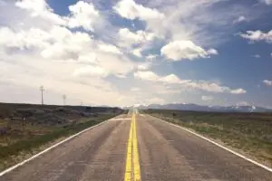 A straight single carriageway road, leading far into the distance, with mountains on the horizon. The route is clear, the sun is shining, and there is nothing on either side of the road.