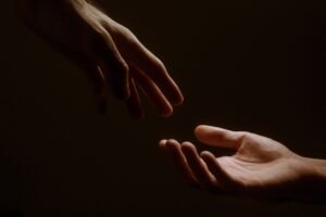 Two hands reaching toward each other in a dark background, fingers nearly touching.