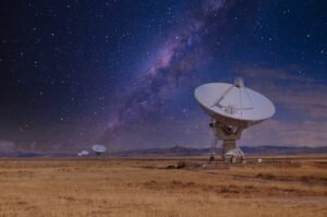 Night sky filled with stars over large radio telescope dishes in a desert plain.
